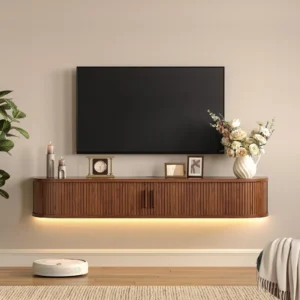 Living room with a wall-mounted TV above a curved wooden console, a vase of pale flowers, plant on the left, and a robotic vacuum on the floor.